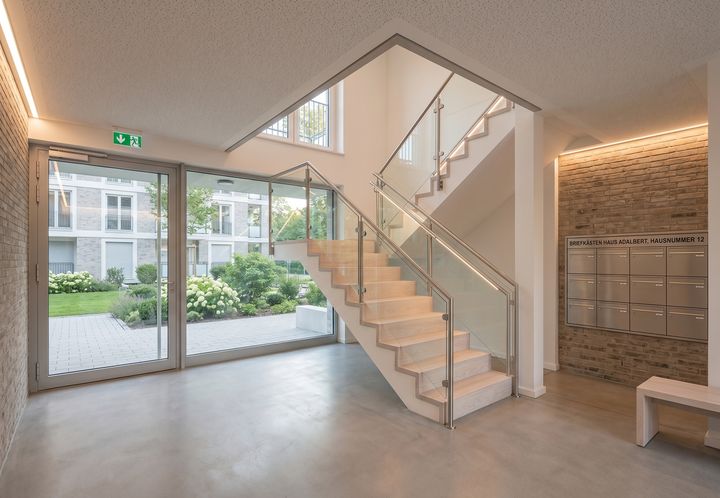 Helles Treppenhaus mit Holztreppe und Glasfront in einem Mehrfamilienhaus (KI-generiertes Symbolbild)